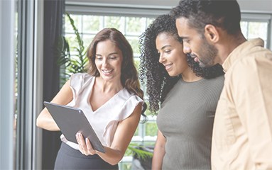 Three coworkers gathered by a window, viewing a tablet and discussing a project.