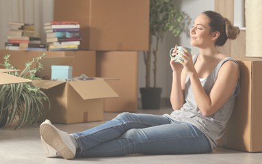 Young woman sits on the floor amid moving boxes, sipping from a mug and taking a break during a move.